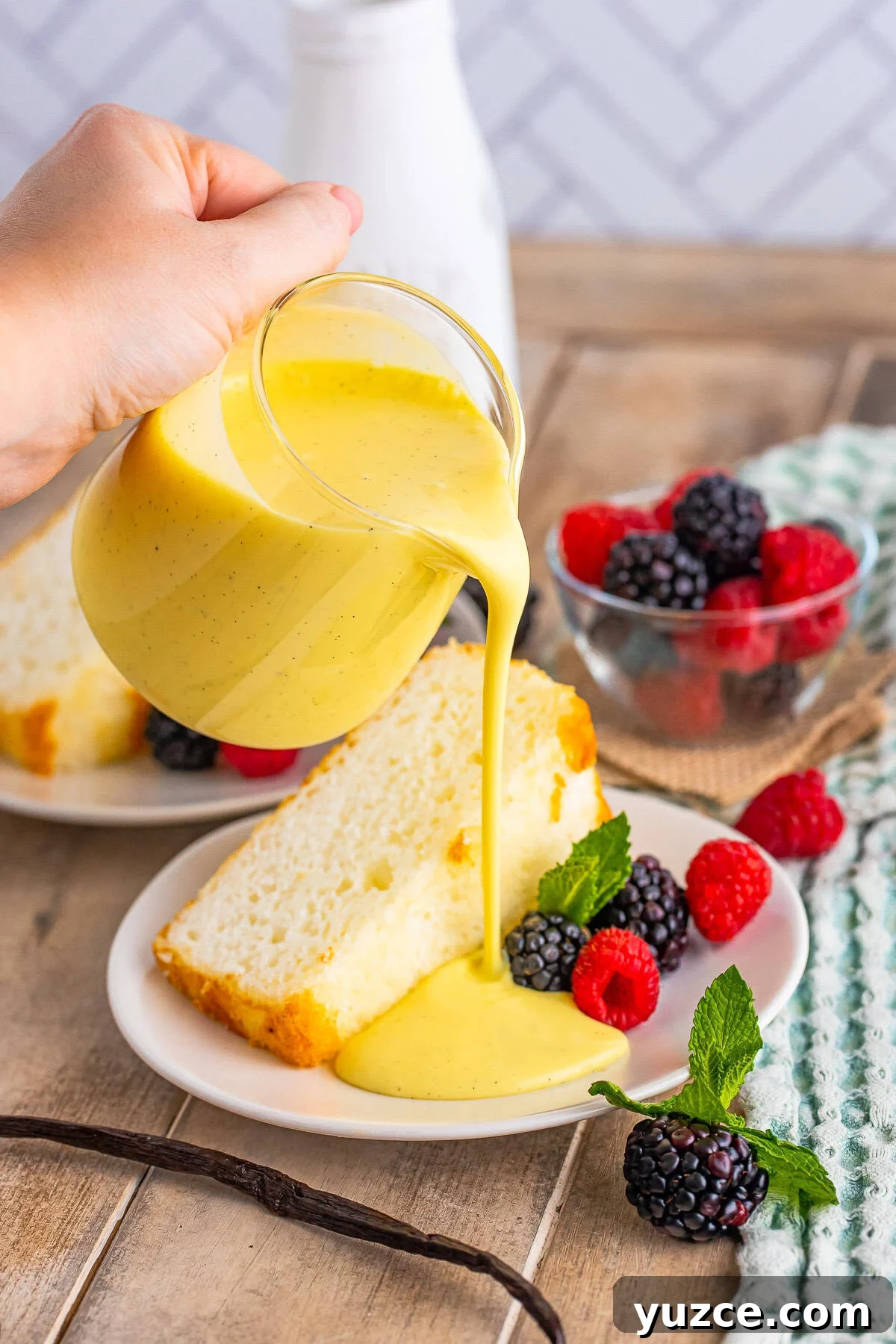 Creme Anglaise Recipe being pour out of a glass with pour spout on to a plate with angel food cake and berries. Served on wooden table top.