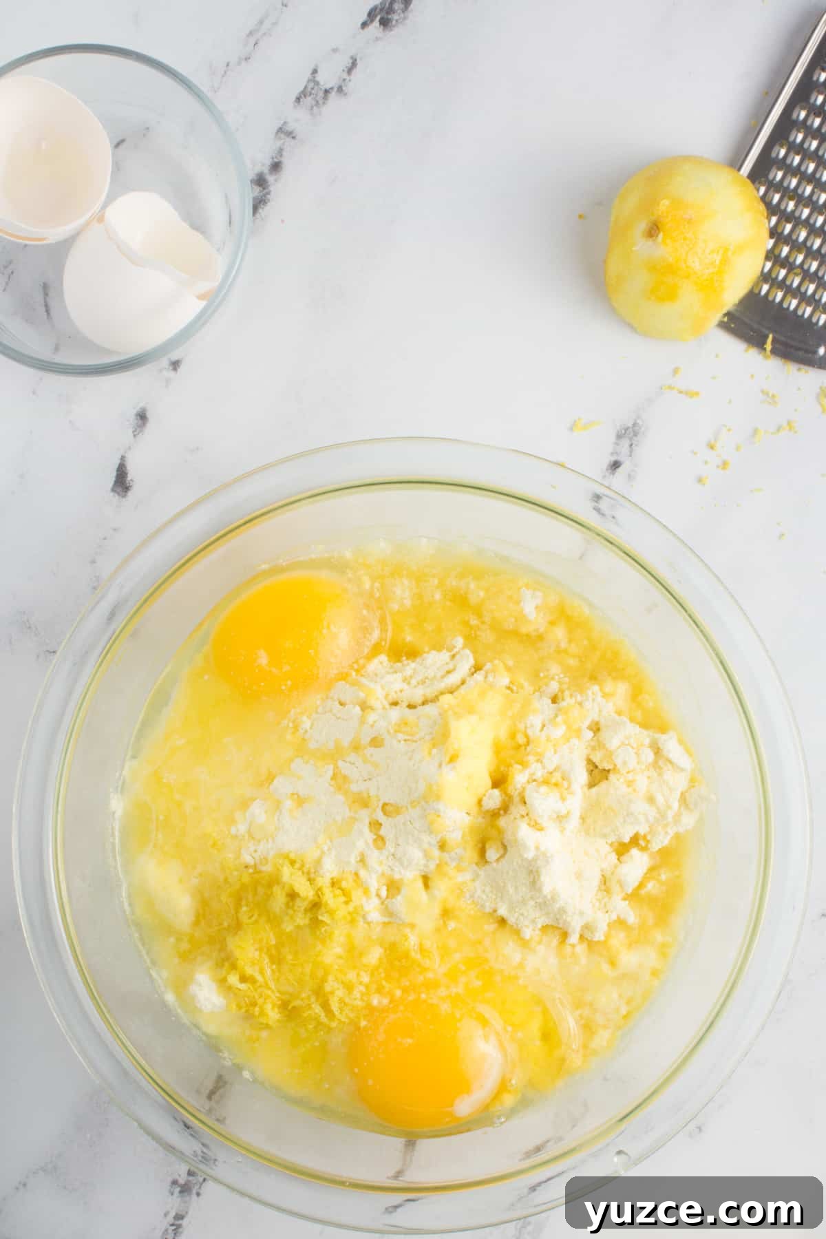 Chewy Lemon Cake Mix Cookies: Quick Citrus Cookie Recipe 5 ingredients for lemon cookies in a bowl partially mixed, sitting on a counter top with lemons, a grater, and empty egg shells around it.