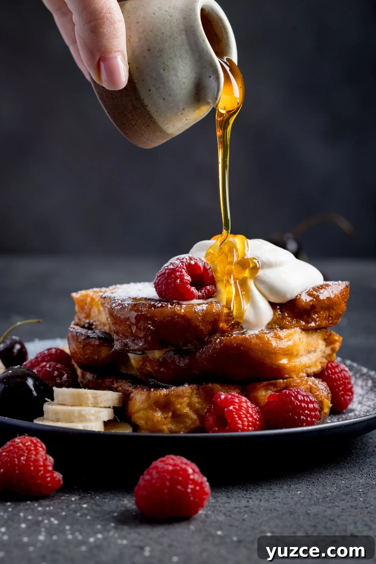 A hand pours syrup onto a stack of caramelised French toast topped with whipped cream, raspberries, cherries, and banana slices on a black plate.