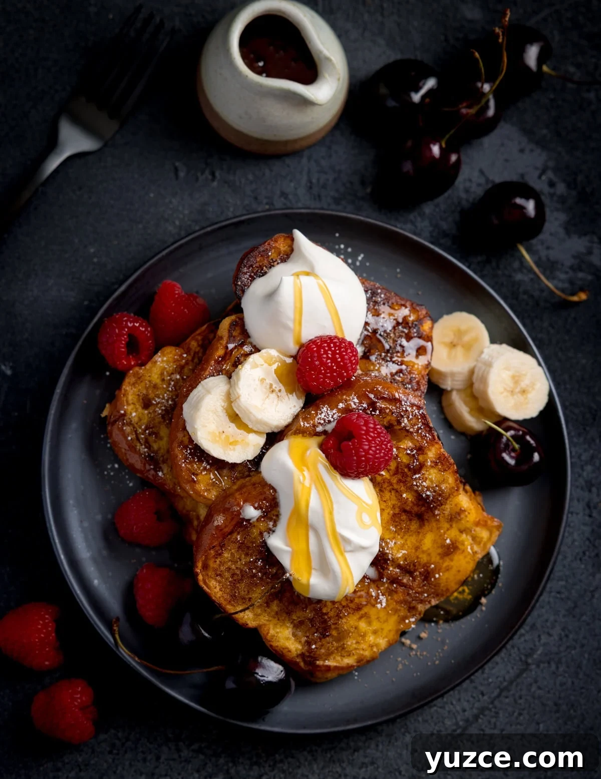 A plate of brioche French toast topped with whipped cream, banana slices, raspberries, cherries, and syrup, with extra fruit and a small jug of syrup nearby.