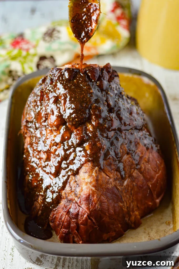 Brown Sugar and Whole Grain Mustard Glazed Baked Ham 7 A bone-in ham with brown sugar glaze being poured over it from a spoon above. It sits in a shallow roasting pan ready to go in the oven.