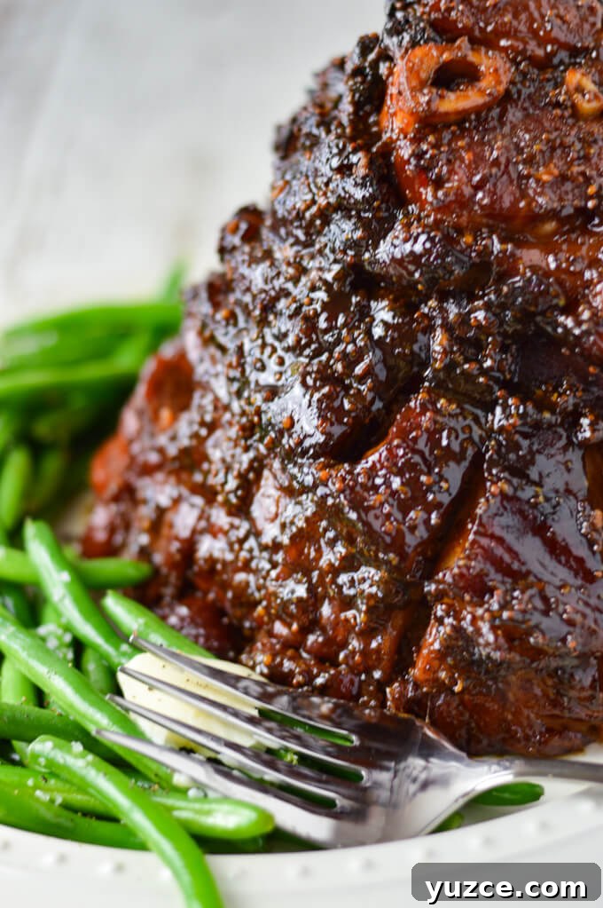 Brown Sugar and Whole Grain Mustard Glazed Baked Ham 8 A close-up view of a cooked glazed ham sitting on a white platter, a large serving fork sits next to it along with a side of green beans.