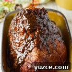 A bone-in ham with brown sugar glaze being poured over it from a spoon above. It sits in a shallow roasting pan ready to go in the oven.