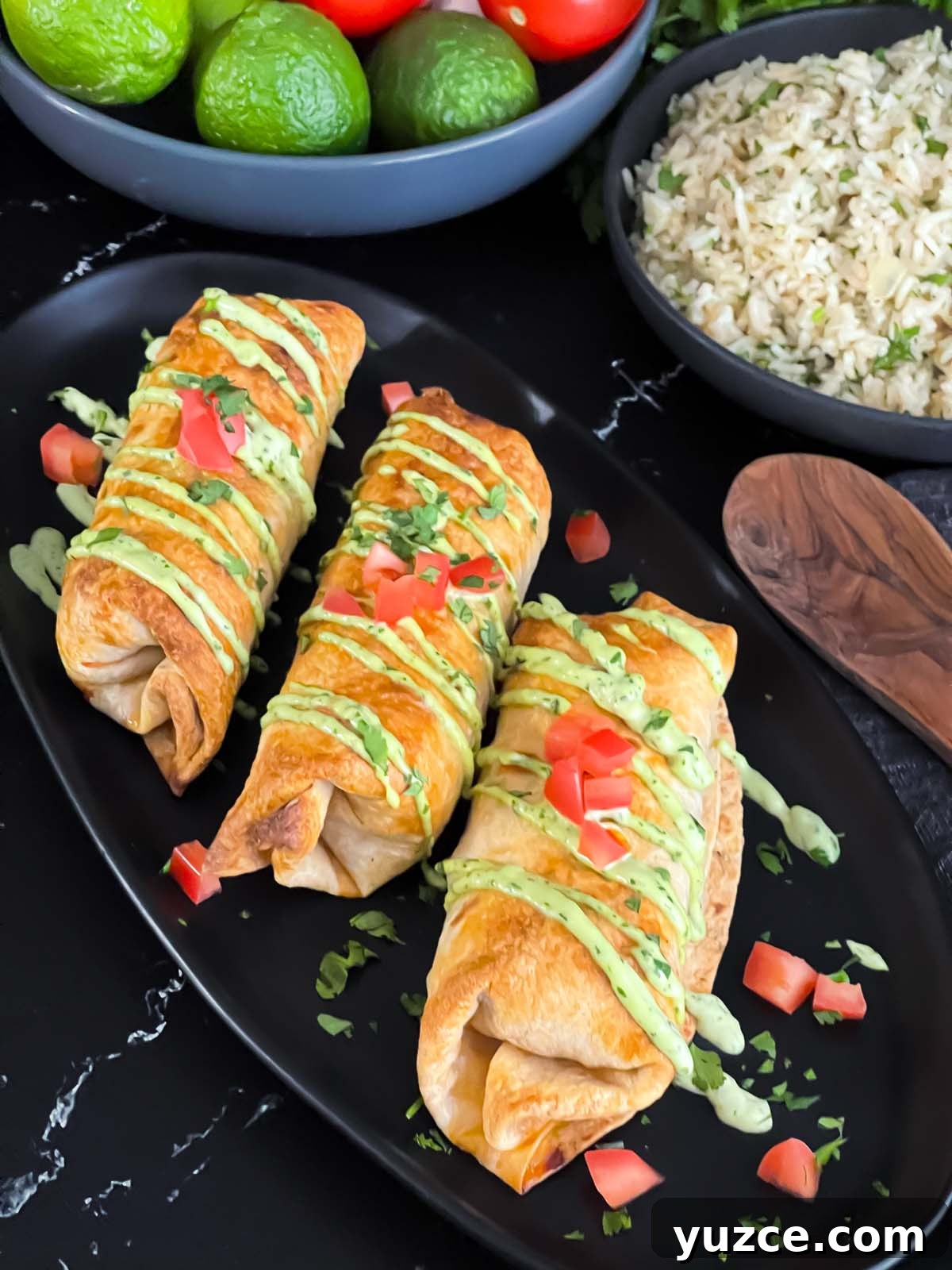 Crispy Baked Chicken Chimichangas with Cheesy Filling 2 Baked chicken chimichangas on a dark oval platter garnished with avocado crema, diced tomatoes and cilantro.