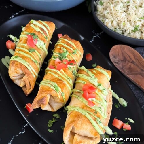 Baked chicken chimichangas on a dark oval platter garnished with avocado crema, diced tomatoes and cilantro.