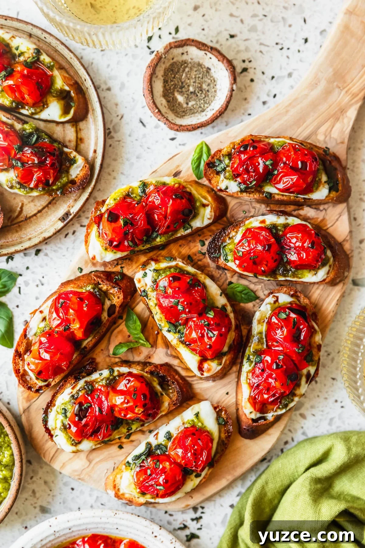 Baked Caprese Crostini with Fresh Basil Pesto 2 A wood board with baked Caprese crostini on a white terrazzo counter next to glasses of white wine, a green linen, and white bowls of tomatoes and pepper.