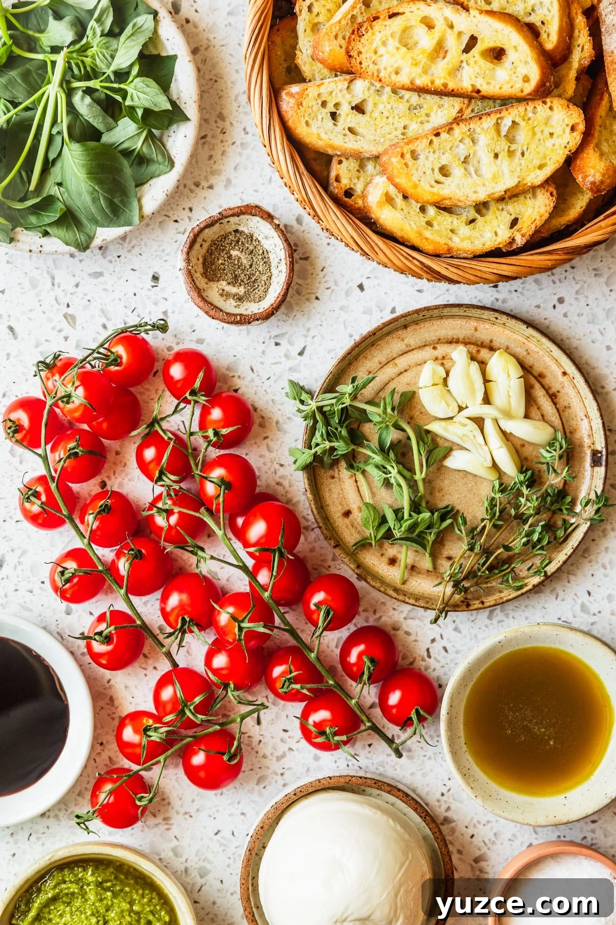 Baked Caprese Crostini with Fresh Basil Pesto 3 White and brown bowls of garlic, herbs, oil, vinegar, crackers, basil, mozzarella, and pesto on a white terrazzo counter next to tomatoes.
