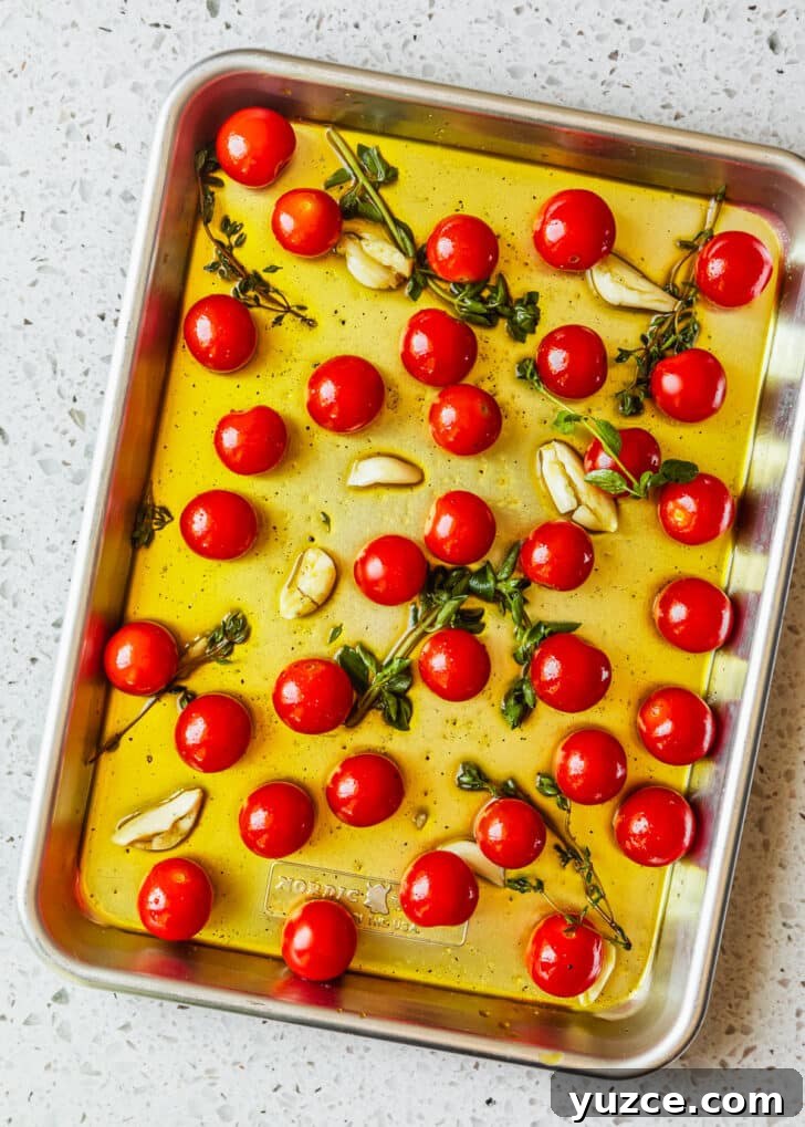 Baked Caprese Crostini with Fresh Basil Pesto 5 A silver sheet pan of cherry tomatoes tossed with oil, garlic, and herbs on a white counter.