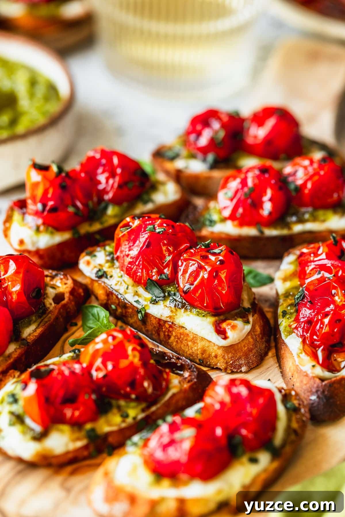 Baked Caprese Crostini with Fresh Basil Pesto 10 A wood board of Caprese crostini on a white counter next to glasses of white wine and a white bowl of pesto.