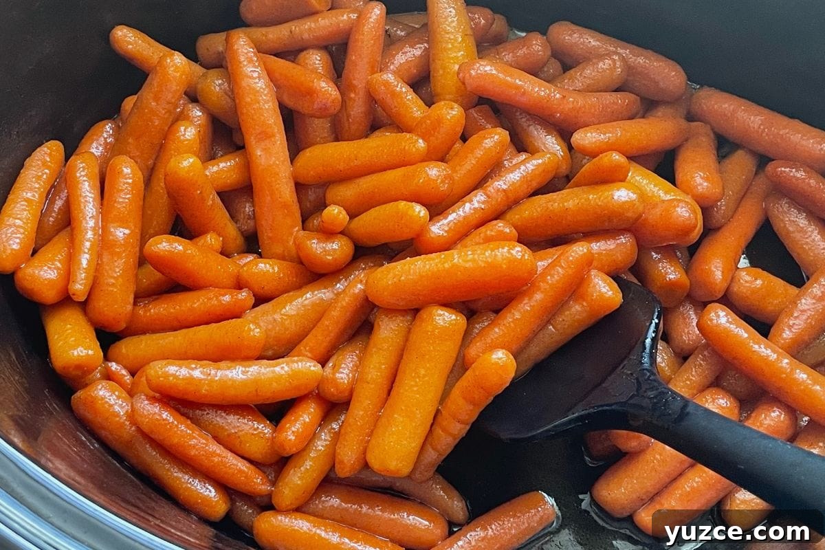 13 Crockpot Thanksgiving Recipes to Save Oven Space 7 A close-up view of shiny glazed baby carrots in a black cooking pot with a black spatula.
