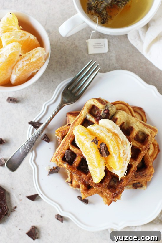 Recipe for orange and dark chocolate waffles. Made with white whole wheat flour and coconut oil! Filled with orange zest and chopped dark chocolate! Several Orange and Dark Chocolate Waffles stacked on a white plate with a cup of tea to the side.