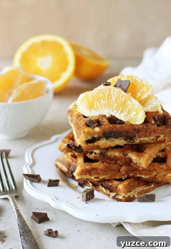 Recipe for orange and dark chocolate waffles. Made with white whole wheat flour and coconut oil! Filled with orange zest and chopped dark chocolate! A white plate stacked with four Orange Waffles.