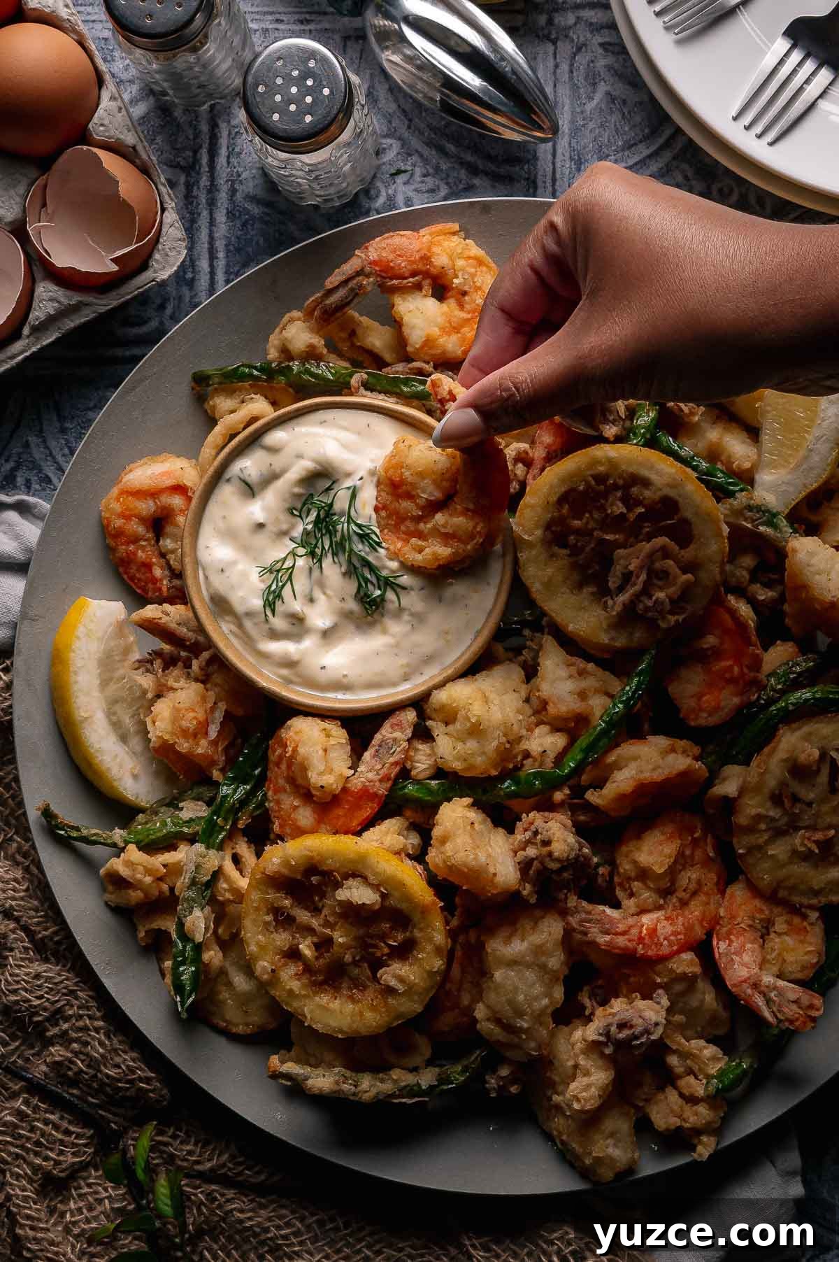 Crispy Fritto Misto with Lemon-Dill Tartar Sauce 7 A hand dips a fried shrimp into a bowl of creamy dipping sauce on a platter with fried shrimp, vegetables, lemon slices, and garnishes.