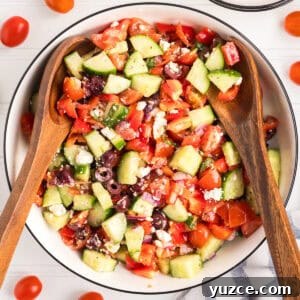 A large bowl of colorful Greek salad, filled with chopped cucumbers, cherry tomatoes, red bell peppers, kalamata olives, red onion, crumbled feta, and herbs. Two wooden salad servers rest on the salad.