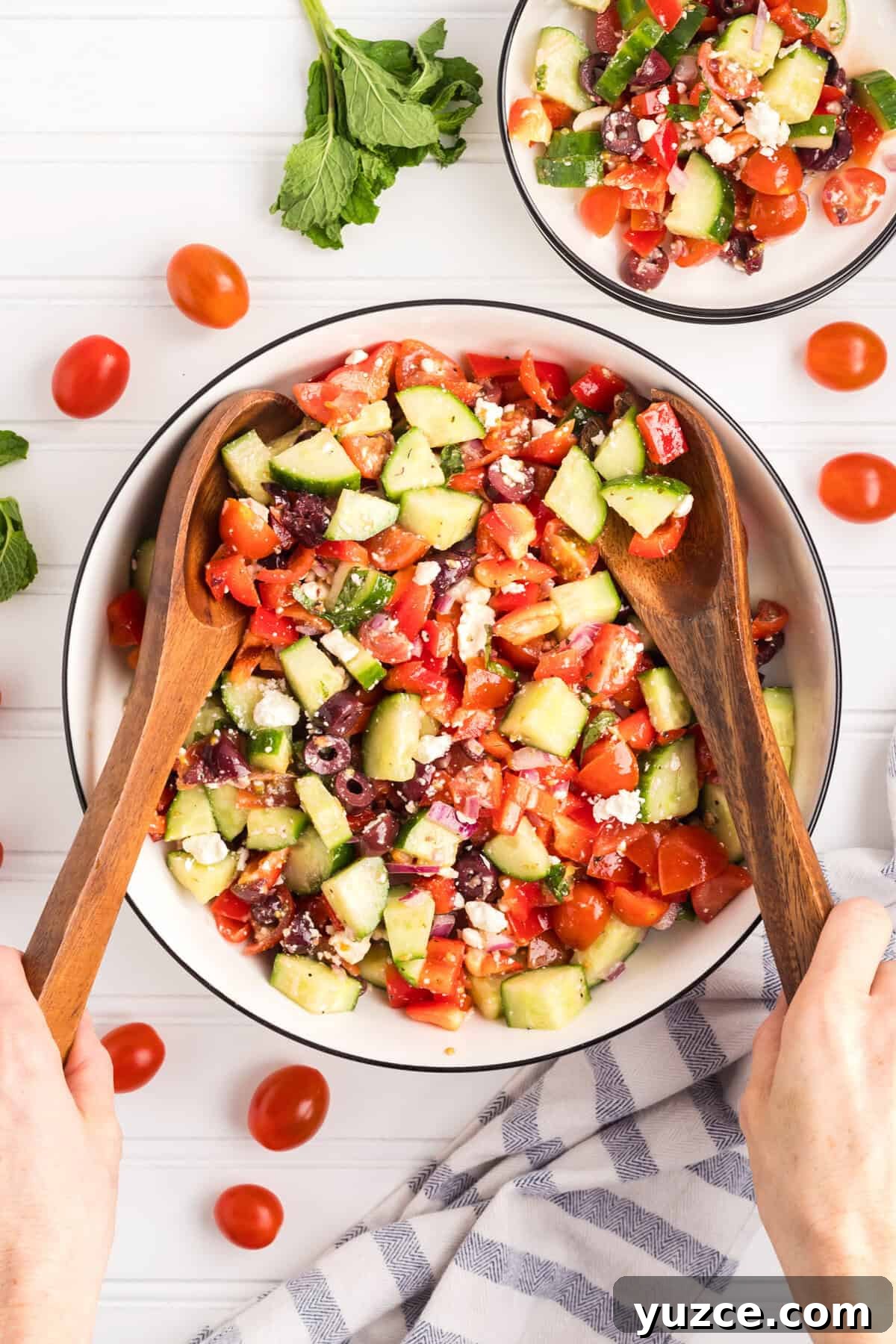 A large bowl of freshly prepared Greek salad with hands holding wooden salad servers, mixing the ingredients.