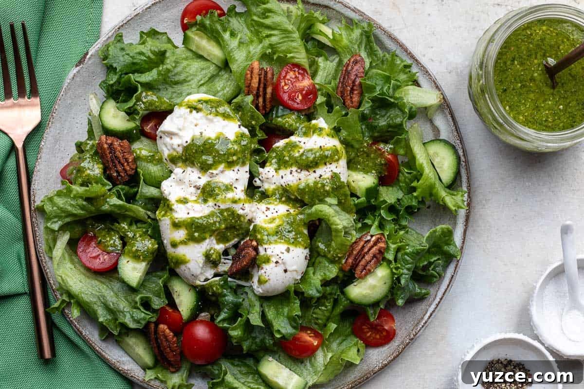 Creamy Pesto Salad Dressing Recipe for Fresh Greens 5 plate with salad greens, cucumber, tomatoes, pecans, burrata cheese, drizzled with pesto dressing next to fork, napkin, and jar of pesto dressing.
